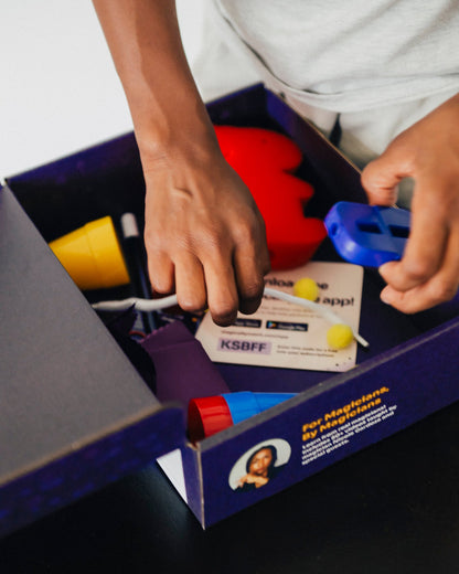 Children’s hands exploring an open Magically Me Kids Magic Kit box, lifting custom playing cards, cup & balls, and disappearing coin—an inclusive beginner‑to‑pro magic set with built‑in SEL learning prompts (ships in 1–2 weeks)